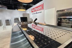 Zbliżenie błyszczącego ekspresu do kawy La Pavoni wystawionego na stoisku targowym La Pavoni, z górnym oświetleniem i czerwonym znakiem La Pavoni na tylnej ścianie. projekt i nadzór Tatjana Kobuszewska trksystem.com