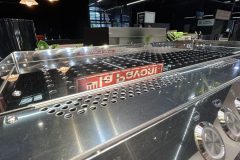 Błyszczący ekspres do kawy ze stali nierdzewnej z pokrętłami i otworami wentylacyjnymi jest wyświetlany pod czerwono-białym znakiem la Pavoni w dobrze oświetlonym, nowoczesnym salonie. Znak odbija się na powierzchni ekspresu. projekt i nadzór Tatjana Kobuszewska trksystem.com