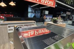 Błyszczący ekspres do kawy La Pavoni jest prezentowany na stoisku wystawowym z dużym czerwonym znakiem La Pavoni nad głową. W tle, przy nowoczesnym oświetleniu, widoczni są ludzie i lady wystawowe. projekt i nadzór Tatjana Kobuszewska trksystem.com