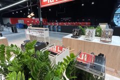 Nowoczesny salon wystawowy prezentuje różne ekspresy do kawy na drewnianych ladach, z zielonymi roślinami na pierwszym planie i dużym czerwonym znakiem la Pavoni zwisającym z sufitu. projekt i nadzór Tatjana Kobuszewska trksystem.com