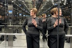 Tatjana Kobuszewska stoi w lustrzanej windzie, tworząc wiele odbić. Trzyma telefon i patrzy w lewo, podczas gdy podłoga i sufit również odbijają jej wizerunek, zwielokrotniając jej wygląd w całej przestrzeni. projekt i nadzór Tatjana Kobuszewska trksystem.com