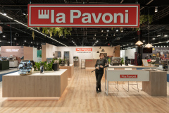 Tatjana Kobuszewska stoi na stoisku la Pavoni, otoczona nowoczesnymi ekspresami do kawy, roślinami i ladami wystawowymi pod dużym wiszącym szyldem marki. Stoisko wyróżnia się drewnianą podłogą i współczesną aranżacją. projekt i nadzór Tatjana Kobuszewska trksystem.com