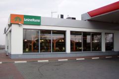 Przydrożna restauracja z dużymi oknami i napisem „Leśne Runo” nad wejściem, obok zadaszenia stacji benzynowej. Biały budynek z zielonymi akcentami odzwierciedla doświadczenie projekt i nadzór Tatjana Kobuszewska trksystem.com. projekt i nadzór Tatjana Kobuszewska trksystem.com
