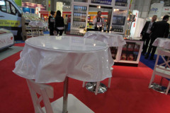Biały stół z plastikową osłoną i białe krzesło na czerwonym dywanie w stoisku targowym Makro CC podczas targów Eurogastro, z półkami ekspozycyjnymi i ludźmi w tle. projekt i nadzór Tatjana Kobuszewska trksystem.com