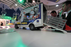Miniaturowy model samochodu dostawczego z brandingiem makro jest wystawiony na stole w stoisku targowym Makro CC, z wizerunkami szefów kuchni na boku i małą rampą załadunkową wysuniętą z tyłu podczas targów Eurogastro. projekt i nadzór Tatjana Kobuszewska trksystem.com