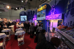 Ludzie gromadzą się w krytym stoisku targowym Makro CC podczas targów Eurogastro, z podświetlanymi wystawami i produktami spożywczymi prezentowanymi na ladach. projekt i nadzór Tatjana Kobuszewska trksystem.com