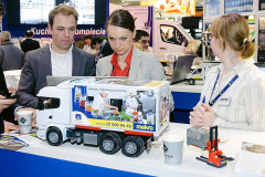 Trzy osoby oglądają szczegółowy model ciężarówki z reklamą Makro na stoisku targowym Makro CC podczas targów Eurogastro, w otoczeniu materiałów promocyjnych i miniaturowego podnośnika paletowego. projekt i nadzór Tatjana Kobuszewska trksystem.com