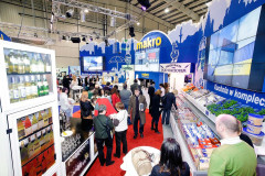 Ludzie gromadzą się na targach Eurogastro wokół stoiska targowego Makro CC, na którym znajdują się półki z produktami, lodówka z napojami i branding Makro na niebiesko-białym wyświetlaczu. projekt i nadzór Tatjana Kobuszewska trksystem.com