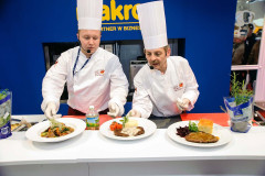 Dwóch kucharzy w białych uniformach i czapkach przygotowuje i prezentuje trzy dania na blacie w stoisku targowym Makro CC podczas targów Eurogastro, z różnymi ziołami i składnikami widocznymi w pobliżu. projekt i nadzór Tatjana Kobuszewska trksystem.com