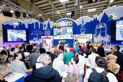 Tłum gromadzi się w stoisku targowym Makro CC, z grafiką żywności i ekranami cyfrowymi, podczas targi Eurogastro w przestronnej, dobrze oświetlonej hali eventowej. projekt i nadzór Tatjana Kobuszewska trksystem.com