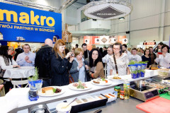 Grupa osób obserwuje i fotografuje dania na talerzach podczas pokazu gotowania podczas targi Eurogastro, z ruchliwym stoiskiem targowym Makro CC widocznym w tle. projekt i nadzór Tatjana Kobuszewska trksystem.com
