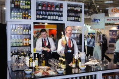 W stoisku targowym Makro CC podczas targów Eurogastro dwóch mężczyzn w mundurach stoi za barem z winami - jeden nalewa wino, drugi trzyma butelkę i mówi do zestawu słuchawkowego - a butelki i kieliszki są starannie ułożone na ladzie. projekt i nadzór Tatjana Kobuszewska trksystem.com