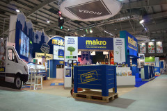 Stoisko targowe Makro CC w hali wystawienniczej zawiera markowe ekspozytory, produkty, grafikę samochodu dostawczego i niebieską skrzynię paletową na pierwszym planie podczas targów Eurogastro. projekt i nadzór Tatjana Kobuszewska trksystem.com