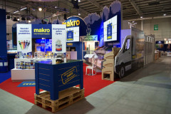 Stoisko targowe Makro CC na targach Eurogastro z firmowymi oznaczeniami, ekspozycją produktów, niebieskim kontuarem i grafiką vana, ustawione w dużej hali wystawowej z czerwoną wykładziną. projekt i nadzór Tatjana Kobuszewska trksystem.com