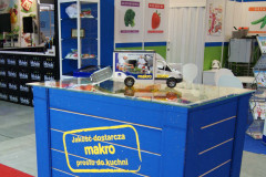 Niebieska lada wystawowa z żółtym logo Makro, samochód dostawczy z zabawkami, mała plastikowa skrzynka i broszury w stoisku targowym Makro CC podczas targów Eurogastro, w tle plakaty spożywcze. projekt i nadzór Tatjana Kobuszewska trksystem.com