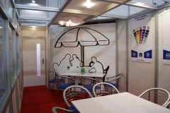 Mała strefa spotkań z metalowymi krzesłami i stołami, fototapetą przedstawiającą ludzi pod parasolem i plakatami informacyjnymi na prawej ścianie - idealna do dyskusji w stoisku targowym Makro CC podczas targów Eurogastro. projekt i nadzór Tatjana Kobuszewska trksystem.com