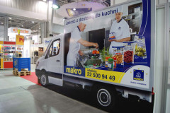 Samochód dostawczy z dużą reklamą przedstawiającą kucharzy przygotowujących jedzenie, z logo Makro i danymi kontaktowymi, jest zaparkowany wewnątrz krytej hali targowej na targach Eurogastro w pobliżu stoiska targowego Makro CC. projekt i nadzór Tatjana Kobuszewska trksystem.com