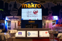 Kryte stoisko gastronomiczne na targach Eurogastro ze stoiskiem targowym Makro CC, dwiema ladami z napisami GregorBurger i Restauracja Pod Kaczorem oraz dużym ekranem cyfrowym wyświetlającym QMP-Dobra Wołowina. projekt i nadzór Tatjana Kobuszewska trksystem.com