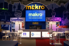 Stoisko targowe Makro CC w targi Eurogastro to podświetlane szyldy, stoliki z białymi nakryciami oraz cyfrowe ekrany z napisami "Makro" i Twój Partner w Biznesie. projekt i nadzór Tatjana Kobuszewska trksystem.com