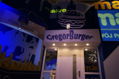 Wejście do restauracji z napisem GregorBurger pod neonową ikoną burgera, otoczone jaskrawymi reklamami i grafikami ściennymi, przypominającymi żywe wyświetlacze w targi Eurogastro lub stoisko targowe Makro CC. projekt i nadzór Tatjana Kobuszewska trksystem.com