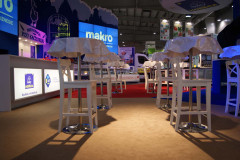 Białe stołki barowe i wysokie stoły pokryte tworzywem sztucznym stoją na wyłożonej wykładziną podłodze na wystawie targi Eurogastro, prezentowanej jako część podświetlanego stoiska targowego Makro CC. projekt i nadzór Tatjana Kobuszewska trksystem.com