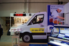 Mężczyzna siedzi wewnątrz białej furgonetki wystawowej w krytym stoisku targowym Makro CC podczas targów Eurogastro, a w tle widoczne są znaki promocyjne i ekspozycje produktów. projekt i nadzór Tatjana Kobuszewska trksystem.com