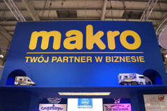 Duży niebiesko-żółty szyld Makro z hasłem Twój partner w biznesie jest wyświetlany nad miniaturowymi modelami ciężarówek i reklamami marek spożywczych na targach Eurogastro na stoisku targowym Makro CC. projekt i nadzór Tatjana Kobuszewska trksystem.com