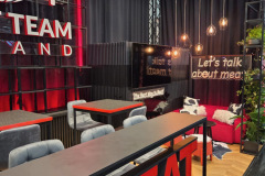 Nowoczesne stoisko wystawiennicze z odważnym czerwonym oznakowaniem "MEAT TEAM", czarnymi ścianami, neonem z napisem "Porozmawiajmy o mięsie", wiszącymi światłami, eleganckimi stołami z szarymi krzesłami, dekoracyjnymi roślinami i przytulnym kącikiem z poduszkami i dywanem. projekt i nadzór Tatjana Kobuszewska trksystem.com