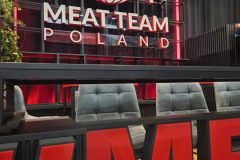 Duży napis "MEAT TEAM POLAND" z kulą ziemską, widelcem i nożem nad nim. Znak jest podświetlony na czerwono. Z przodu znajdują się nowoczesne szare krzesła i częściowy widok na duże czerwone litery tworzące ME. Wygląda to na stoisko wystawowe lub przestrzeń eventową. projekt i nadzór Tatjana Kobuszewska trksystem.com