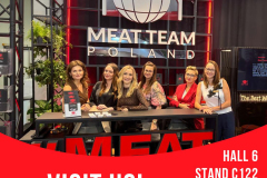 Sześć kobiet stoi uśmiechniętych za ladą na stoisku targowym Meat Team Poland. Na ścianie za nimi widnieje duże logo z kulą ziemską, widelcem i nożem. Stoisko znajduje się w hali 6, stoisko C122 na targach Anuga. Czerwone akcenty i pogrubiony tekst "Odwiedź nas" znajdują się na dole. projekt i nadzór Tatjana Kobuszewska trksystem.com
