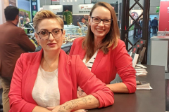 Dwie kobiety w czerwonych marynarkach i okularach stoją uśmiechnięte na stoisku targowym. Jedna opiera się o ladę z wytatuowanymi ramionami, druga stoi za nią. Na blacie stoiska znajduje się miska wypełniona złożonymi papierami. W tle widoczne są wystawy targowe i ludzie. projekt i nadzór Tatjana Kobuszewska trksystem.com