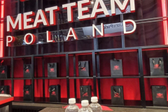 Stół z trzema butelkami wody i czarnym notesem na spirali przed dużym podświetlanym napisem MEAT TEAM POLAND na czerwono-czarnej ścianie. Znak przedstawia kulę ziemską z widelcem i nożem, a małe logo Meat Team Poland zdobi półki w tle. projekt i nadzór Tatjana Kobuszewska trksystem.com