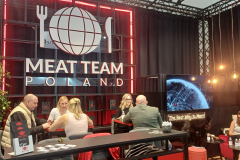Stoisko targowe dla Meat Team Poland przedstawia ludzi siedzących przy stołach i dyskutujących. Tło ma duże logo kuli ziemskiej z czerwonym neonowym oświetleniem. Telewizor wyświetla cyfrową grafikę. Wiszące światła i markowe materiały są widoczne w nowoczesnej aranżacji. projekt i nadzór Tatjana Kobuszewska trksystem.com