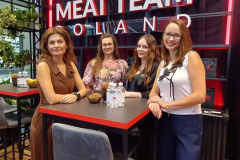 Cztery kobiety stoją i siedzą uśmiechnięte przy wysokim stole przed dużym czerwono-białym napisem MEAT TEAM POLAND z logo kuli ziemskiej, widelca i noża. W nowoczesnym otoczeniu znajdują się rośliny, napoje na stole i czarne metalowe tło. projekt i nadzór Tatjana Kobuszewska trksystem.com