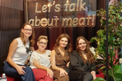 Cztery kobiety siedzą na czerwonej kanapie w przytulnym, wypełnionym roślinami pokoju z wiszącymi lampami i czarną zasłoną. Za nimi znajduje się neon z napisem "Porozmawiajmy o mięsie". Przed nimi stoi mały stolik z notatnikami i napojami. projekt i nadzór Tatjana Kobuszewska trksystem.com