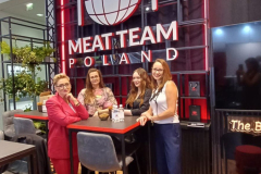 Cztery kobiety stoją i uśmiechają się wokół wysokiego czarnego stołu z napojami. Za nimi znajduje się duży podświetlany napis Meat Team Poland z logo kuli ziemskiej, widelca i noża. Stoisko ma nowoczesny design z czarno-czerwonymi kolorami i drewnianą podłogą. projekt i nadzór Tatjana Kobuszewska trksystem.com