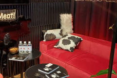 Przytulny salon z czerwoną sofą, poduszkami z krowim nadrukiem, napojami i przekąskami na okrągłym czarnym stole oraz książką. Pod spodem leży dywan ze sztucznej skóry bydlęcej. Powyżej wiszą ciepłe światła, a neon z napisem "Porozmawiajmy o mięsie" uzupełniają ciemne zasłony i panele ścienne. projekt i nadzór Tatjana Kobuszewska trksystem.com