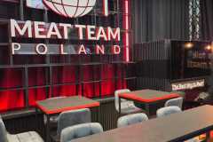 Nowoczesne wnętrze restauracji z dużym podświetlanym napisem "MEAT TEAM POLAND" z kulą ziemską, widelcem i nożem. Przestrzeń ma czarne ściany, czerwone akcenty, szare wyściełane krzesła i stoły, tworząc elegancką i zachęcającą atmosferę. projekt i nadzór Tatjana Kobuszewska trksystem.com