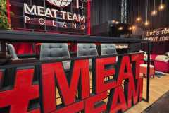 Nowoczesne stoisko z napisem "MEAT TEAM POLAND", czerwonymi neonami, dużym logo w kształcie kuli ziemskiej i naczyniami na tylnej ścianie. Z przodu pogrubione czerwone litery "MEAT TEAM". Są też stołki barowe, wiszące lampy i przytulna strefa wypoczynkowa z roślinami. projekt i nadzór Tatjana Kobuszewska trksystem.com
