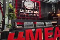 Nowoczesne stoisko targowe dla Meat Team Poland charakteryzuje się odważnym czerwono-czarnym designem, z dużym logo kuli ziemskiej, widelca i noża na tylnej ścianie. Szare krzesła stoją za ladą, a na pierwszym planie znajduje się gigantyczny czerwony znak hashtagu. projekt i nadzór Tatjana Kobuszewska trksystem.com
