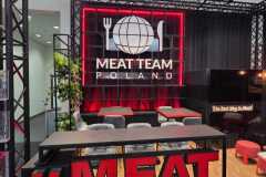 Nowoczesne stoisko targowe ma odważny czerwono-czarny wystrój. Z przodu znajdują się duże czerwone litery 3D z napisem "MEAT TEAM". Z tyłu widnieje kula ziemska z widelcem i nożem oraz napis "MEAT TEAM POLAND". W tle znajdują się stoły, stołki i rośliny. projekt i nadzór Tatjana Kobuszewska trksystem.com