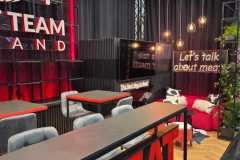 Nowoczesne stoisko targowe posiada oznakowanie "MEAT TEAM" z pogrubionymi czerwonymi literami, szare krzesła przy stołach, czarne ściany z czerwonymi akcentami, wiszące lampy wiszące i neon z napisem "Porozmawiajmy o mięsie". Całość dopełniają rośliny doniczkowe i przytulny dywan. projekt i nadzór Tatjana Kobuszewska trksystem.com