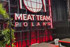 Na ścianie restauracji widnieje napis MEAT TEAM POLAND wykonany dużymi białymi literami. Powyżej znajduje się grafika kuli ziemskiej, widelca i noża oświetlona czerwonymi neonami. Część wypoczynkowa poniżej ma szare krzesła, czarne stoły i jest zaakcentowana zielonymi roślinami. projekt i nadzór Tatjana Kobuszewska trksystem.com