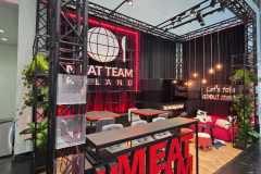 Nowoczesne stoisko wystawiennicze z dużym czerwonym napisem "MEAT TEAM" z przodu. Stoisko ma czarno-czerwony wystrój, rośliny, leżaki i neonowe logo z napisem "MEAT TEAM POLAND". Wiszące światła i napis "Porozmawiajmy o mięsie" znajdują się na tylnej ścianie. projekt i nadzór Tatjana Kobuszewska trksystem.com