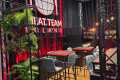 Nowoczesne stoisko z wysokimi stołami i szarymi stołkami barowymi na drewnianej podłodze. Czarne metalowe ramy podtrzymują wiszące rośliny. Podświetlony na czerwono napis "EAT TEAM POLAND" z ikonami widelca, noża i kuli ziemskiej. W rogu wiszą żarówki Edisona, tworząc przytulną atmosferę. projekt i nadzór Tatjana Kobuszewska trksystem.com