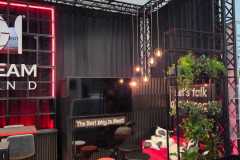 Nowoczesny salon na stoisku eventowym z czerwoną sofą, czarnymi krzesłami, okrągłymi stołami, wiszącymi żarówkami, dywanem w zwierzęcy wzór, zieloną ścianą z roślinami i neonem z napisem "The Best Way to Meet". Przestrzeń zdobią czarne zasłony i metalowe ramy. projekt i nadzór Tatjana Kobuszewska trksystem.com