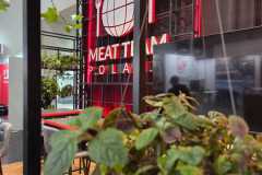 Zbliżenie na zielone rośliny liściaste na pierwszym planie z rozmytym tłem przedstawiającym czerwony neon z napisem MEAT TEAM POLAND z grafiką kuli ziemskiej, widelca i noża. Wydaje się, że jest to wewnętrzne stoisko wystawowe lub targowe. projekt i nadzór Tatjana Kobuszewska trksystem.com