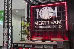 Nowoczesne stoisko wystawowe z czarną metalową ramą, czerwonymi neonami i dużym napisem "MEAT TEAM" z przodu. Z tyłu znajduje się logo przedstawiające widelec, kulę ziemską i nóż z napisem "MEAT TEAM POLAND" pogrubioną białą czcionką. Wewnątrz znajdują się stołki barowe i stoły. projekt i nadzór Tatjana Kobuszewska trksystem.com