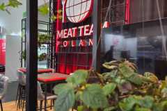 Roślina na pierwszym planie częściowo zasłania nowoczesną budkę z czerwonym neonem z napisem MEAT TEAM POLAND z pogrubionymi literami, z logo kuli ziemskiej i widelca. Stoisko ma wysokie stoły, krzesła i czarne metalowe ramy w stylu industrialnym. projekt i nadzór Tatjana Kobuszewska trksystem.com