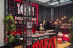 Nowoczesne stoisko wystawiennicze z czarno-czerwonym wystrojem, z oznakowaniem MEAT TEAM POLAND, dużym czerwonym wyświetlaczem MEATTEAM, ladą, zielenią, miejscem do siedzenia, neonami i osobą wewnątrz regulującą element ścienny. Przestrzeń ma stylowy, industrialny wygląd. projekt i nadzór Tatjana Kobuszewska trksystem.com